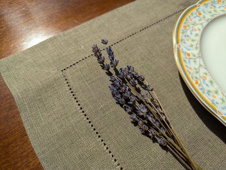 Puede incluir: Un mantel individual de lino beige con un borde decorativo cosido. Una rama de lavanda seca se coloca sobre el mantel, y un plato de porcelana blanca con un dise&ntilde;o floral est&aacute; en el fondo.
