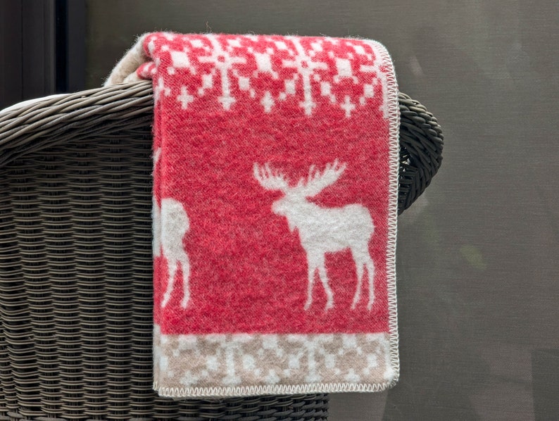 May include: A red and white patterned throw blanket with a white moose design. The blanket is folded and draped over a wicker chair.