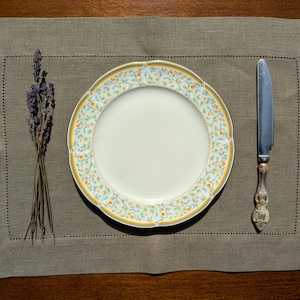 Puede incluir: Un plato de cer&aacute;mica blanca con un patr&oacute;n floral en tonos de amarillo, verde y azul. El plato est&aacute; colocado sobre un mantel individual de lino marr&oacute;n con un cuchillo a la derecha y un ramo de lavanda seca a la izquierda.