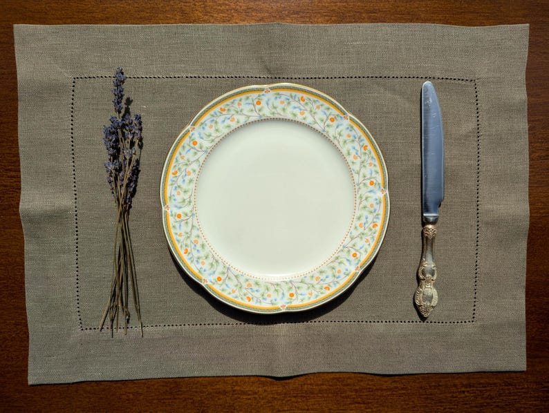 Puede incluir: Un plato de cer&aacute;mica blanca con un patr&oacute;n floral en tonos de amarillo, verde y azul. El plato est&aacute; colocado sobre un mantel individual de lino marr&oacute;n con un cuchillo a la derecha y un ramo de lavanda seca a la izquierda.