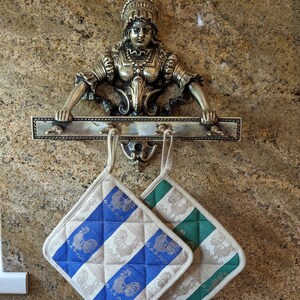 Puede incluir: Dos agarraderas de olla con un patr&oacute;n a cuadros azul y blanco y un patr&oacute;n a cuadros verde y blanco cuelgan de un estante de metal con una figura decorativa de mujer de lat&oacute;n.