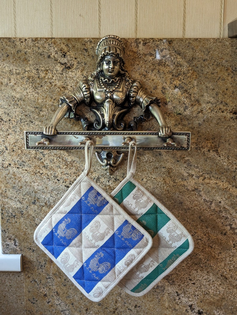 Puede incluir: Dos agarraderas de olla con un patr&oacute;n a cuadros azul y blanco y un patr&oacute;n a cuadros verde y blanco cuelgan de un estante de metal con una figura decorativa de mujer de lat&oacute;n.