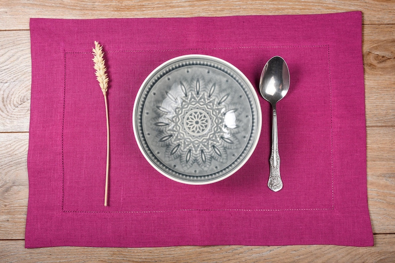 May include: A gray ceramic bowl with a decorative design sits on a pink linen placemat with a silver spoon and a wheat stalk.