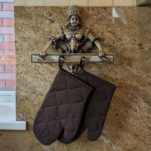 K&ouml;nnte beinhalten: Ein dekorativer Metallst&auml;nder mit einer Frauenfigur, die einen Handtuchhalter h&auml;lt. Zwei braune Ofenhandschuhe h&auml;ngen an dem St&auml;nder.