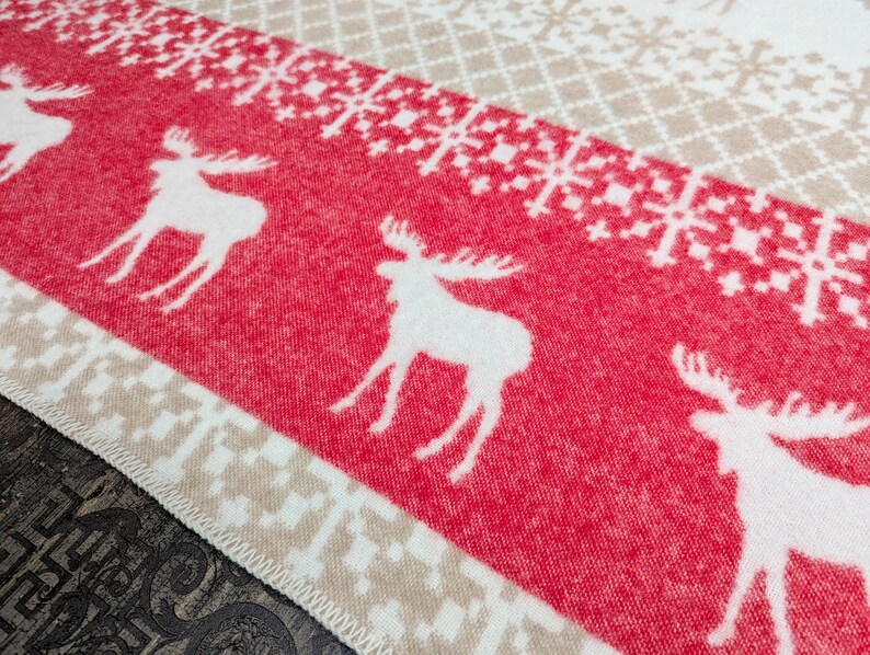 May include: A red and white patterned throw blanket with a white moose design. The blanket has a beige border.