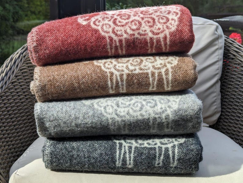 May include: A stack of four wool blankets with a sheep design. The blankets are in shades of red, brown, gray, and black.