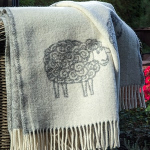 May include: A white throw blanket with a gray sheep design. The blanket has a fringe border and is draped over a wicker chair.