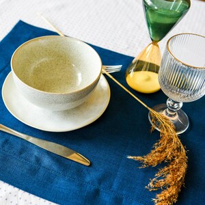 Puede incluir: Un mantel individual azul con un taz&oacute;n blanco, un platillo blanco, un cuchillo plateado, un vaso de agua y un arreglo de flores secas. El mantel individual est&aacute; sobre un mantel blanco con peque&ntilde;os puntos blancos.