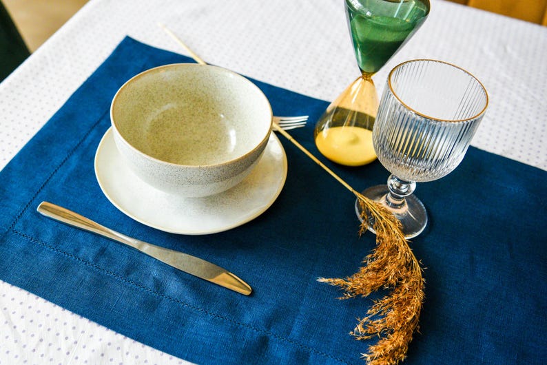 Puede incluir: Un mantel individual azul con un taz&oacute;n blanco, un platillo blanco, un cuchillo plateado, un vaso de agua y un arreglo de flores secas. El mantel individual est&aacute; sobre un mantel blanco con peque&ntilde;os puntos blancos.
