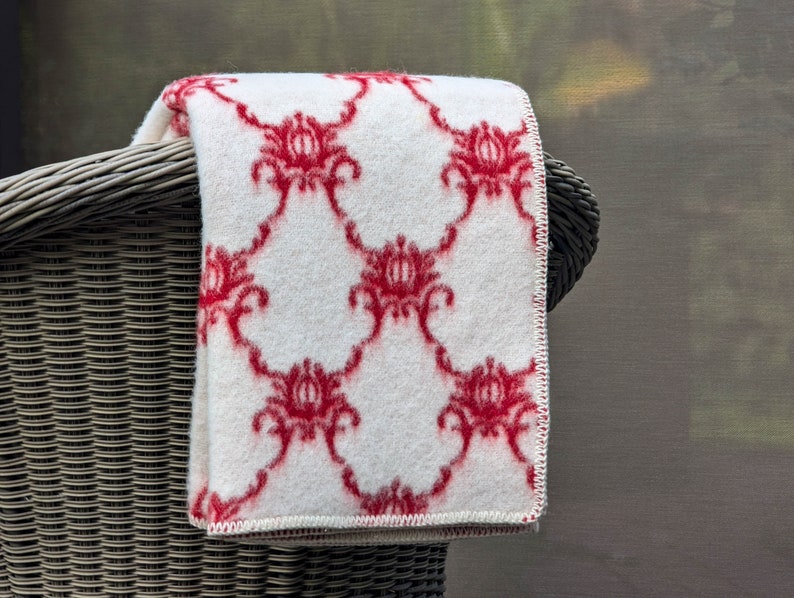 May include: White throw blanket with a red floral pattern. The blanket is folded and draped over a wicker chair.