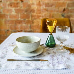 May include: A white ceramic bowl with a saucer, a glass of water, and an hourglass with yellow sand and a green base on a white tablecloth with a floral pattern.