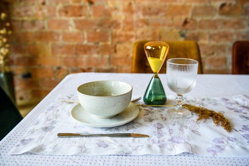 May include: A white ceramic bowl with a saucer, a glass of water, and an hourglass with yellow sand and a green base on a white tablecloth with a floral pattern.
