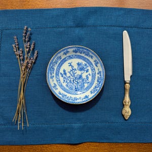 May include: A blue linen placemat with a white and blue patterned plate, a silver knife, and a bunch of lavender.