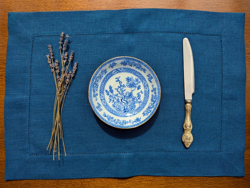May include: A blue linen placemat with a white and blue patterned plate, a silver knife, and a bunch of lavender.