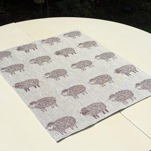 May include: A light grey linen table runner with a repeating pattern of brown sheep. The sheep are stylised and have curly wool.
