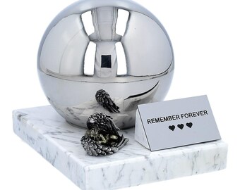 May include: A polished silver sphere-shaped urn with two angel figurines and a marble base. A small white card with the words "REMEMBER FOREVER" and three heart symbols is also present. The urn is designed for memorial purposes.