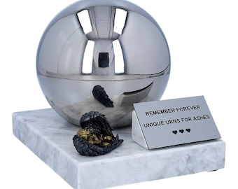 May include: A polished silver urn with a spherical design, resting on a white marble base. A small, gold-toned angel figure with black wings is nestled near the urn. A silver plaque reads "REMEMBER FOREVER UNIQUE URNS FOR ASHES".