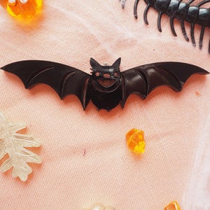 May include: Black bat-shaped pin with a smiling face. The pin is made of plastic and has a shiny finish.