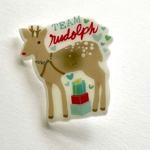 May include: A white acrylic pin with a cartoon illustration of a brown reindeer with a red nose, wearing a harness and pulling a stack of wrapped gifts. The text "TEAM rudolph" is printed above the reindeer with blue and green hearts.