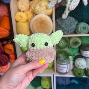 May include: A green and brown crocheted plush toy resembling a popular fictional character. The toy is being held in a hand with yellow nail polish.