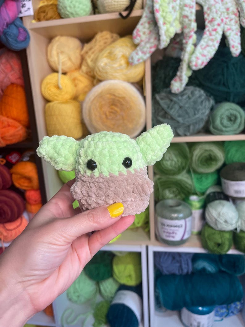 May include: A green and brown crocheted plush toy resembling a popular fictional character. The toy is being held in a hand with yellow nail polish.