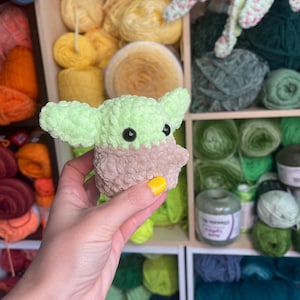 May include: A small green and brown crocheted plush toy resembling a character from the Star Wars franchise. The toy is being held by a hand with yellow nail polish.