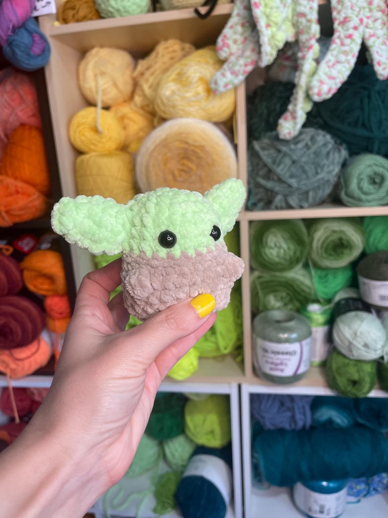 May include: A small green and brown crocheted plush toy resembling a character from the Star Wars franchise. The toy is being held by a hand with yellow nail polish.