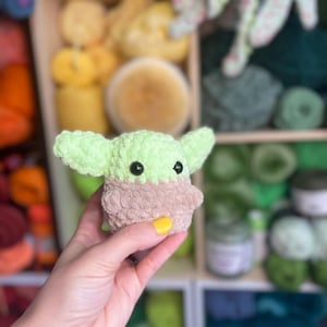 May include: A small green crocheted plush toy resembling a character from the Star Wars franchise. The toy has large black eyes and a brown fuzzy body.