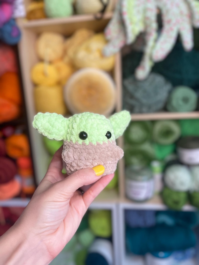 May include: A small green crocheted plush toy resembling a character from the Star Wars franchise. The toy has large black eyes and a brown fuzzy body.