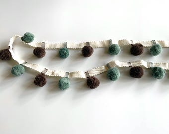 May include: A white trim with brown and green pom poms. The trim is made of a woven fabric with a striped pattern.