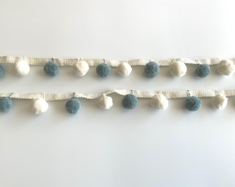 May include: Two rows of pompom trim. Each row has a cream-colored woven base with alternating blue-green and white pom-poms. The pom-poms are evenly spaced along the trim, creating a decorative border.
