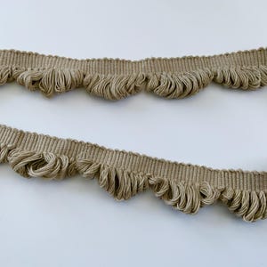 May include: A close-up of beige fabric trim with a scalloped edge and a fringe detail. The trim is made of a woven fabric with a subtle texture.