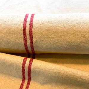 May include: A close-up of a natural linen fabric with two red stripes running down the length of the fabric. The fabric is folded in half, and the red stripes are visible on both sides of the fold.