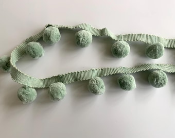 May include: A sage green pom-pom trim with a woven ribbon base. The trim features a row of fluffy pom-poms.