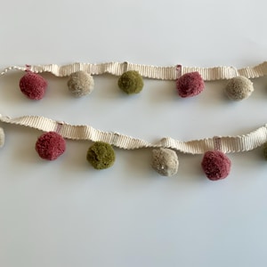May include: Two lengths of decorative trim featuring alternating pom-poms in olive green, burgundy, and beige. The trim is a cream-coloured woven ribbon with a subtle ribbed texture. The pom-poms are evenly spaced along the ribbon.
