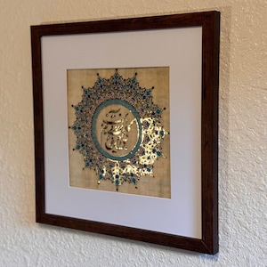 May include: A framed artwork featuring a gold calligraphy design with blue and pink accents on a beige background. The design is surrounded by a circular pattern of intricate details.