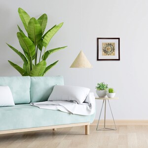 May include: A light blue sofa with white pillows and a throw blanket. A small wooden table with two potted plants sits next to the sofa. A framed print with a circular design hangs on the wall above the table. A light wood pendant lamp hangs above the sofa.