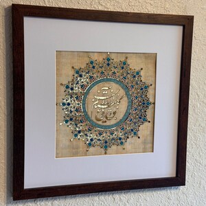 May include: A framed artwork featuring a circular design with intricate gold and blue patterns. The center of the design contains Arabic calligraphy in gold and blue.