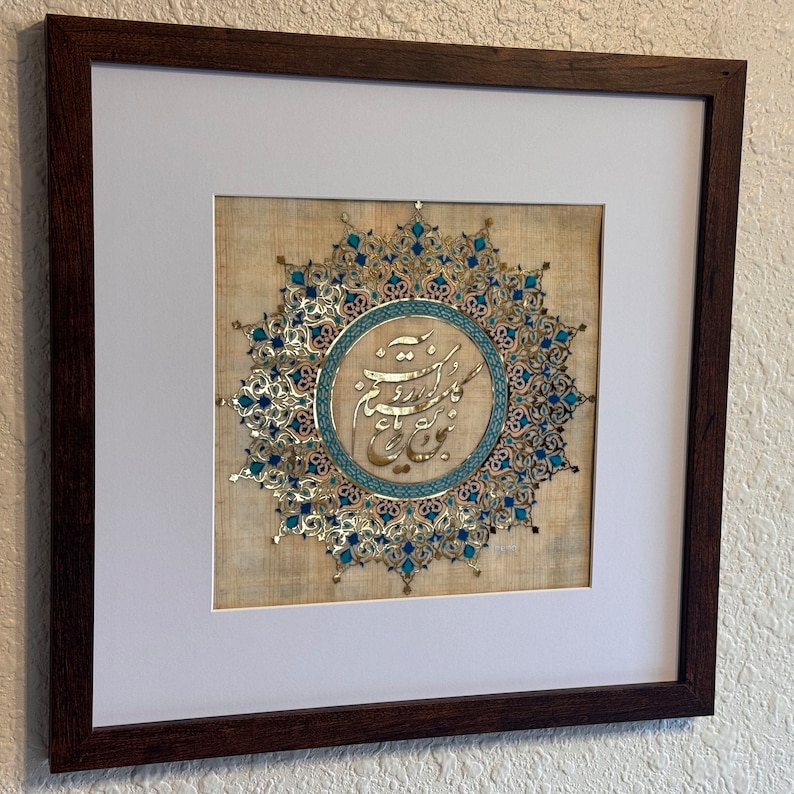 May include: A framed artwork featuring a circular design with intricate gold and blue patterns. The center of the design contains Arabic calligraphy in gold and blue.