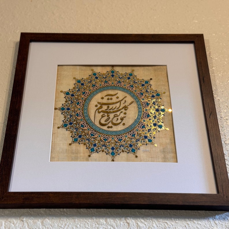 May include: A framed artwork featuring a gold and blue mandala design with Arabic calligraphy in the center. The artwork is on a beige background and is framed in a dark brown wood frame.