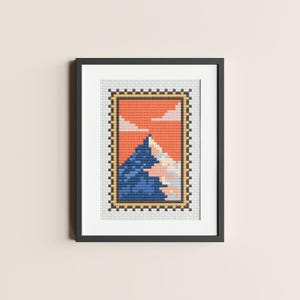 May include: A framed cross-stitch artwork depicting a mountain range with a blue peak and a pink and white snow-capped peak against an orange sunset sky.