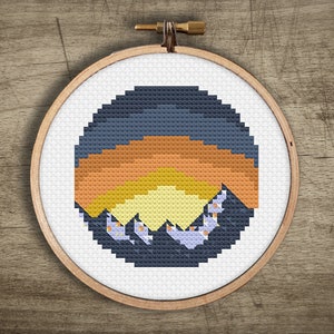 May include: A cross-stitch embroidery hoop with a circular design of a sunset over mountains. The mountains are purple, the sunset is orange, yellow, and light blue, and the sky is dark blue.
