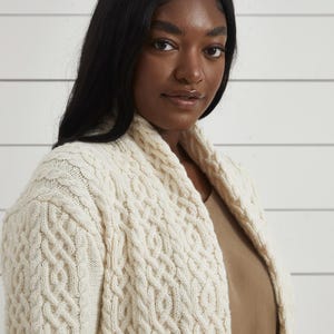 May include: Cream cable knit cardigan with a shawl collar. The sweater has a textured pattern and is made of a soft, warm material. A cozy and stylish outerwear option.