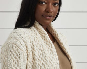 May include: Cream cable knit cardigan with a shawl collar. The sweater has a textured pattern and is made of a soft, warm material. A cozy and stylish outerwear option.