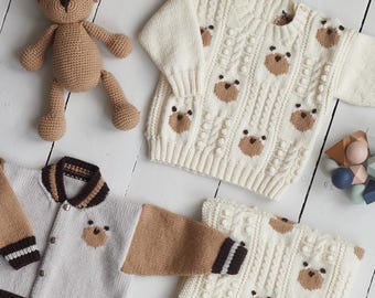 May include: A collection of baby clothing and accessories, including a knitted teddy bear, a hat, a sweater, a jacket, and a blanket. The sweater and blanket feature a repeating bear face pattern. The jacket has brown sleeves and trim.