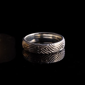 May include: A silver ring with a textured, organic pattern. The ring is reflecting light on a black surface.