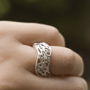 May include: A silver ring with a unique, organic, openwork design. The ring is worn on a finger and has a wavy, asymmetrical shape.