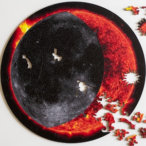 May include: A circular wooden jigsaw puzzle depicting a solar eclipse with the moon covering the sun. The puzzle is partially assembled, with some pieces missing. The puzzle features a detailed image of the sun and moon, with a fiery red and orange color scheme.