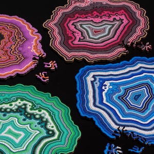 May include: Four colourful wooden jigsaw puzzles with an agate stone design. The puzzles are in shades of purple, pink, blue, and green, with intricate layered patterns. Several puzzle pieces are scattered around the completed puzzles on a black surface.