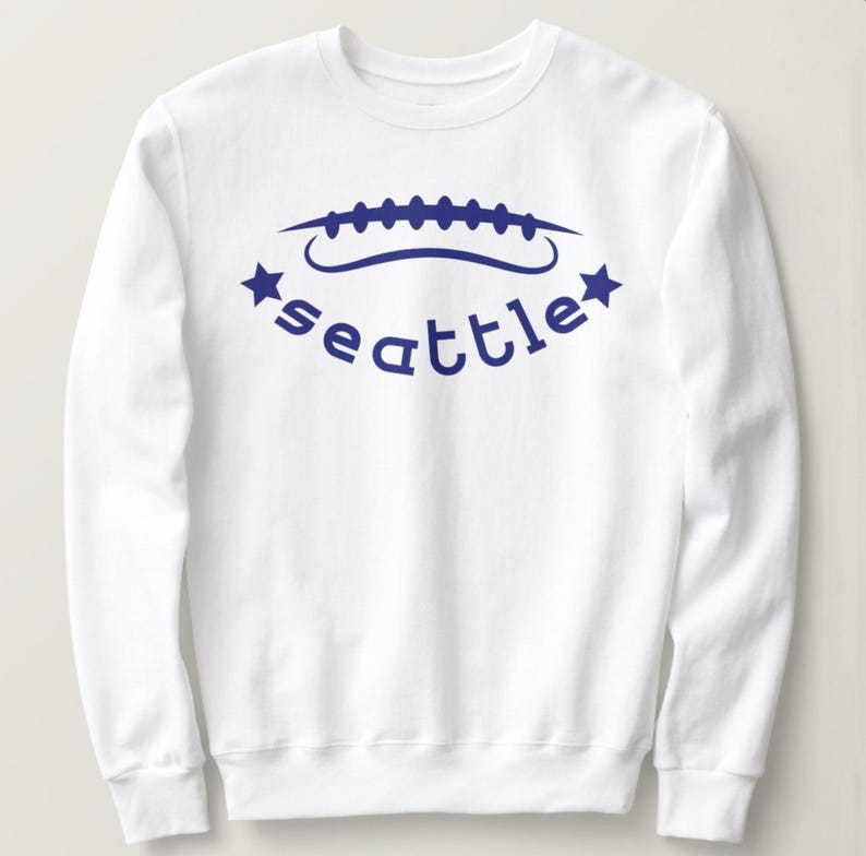 May include: White sweatshirt with a blue football design and the word "Seattle" in a curved font. The design includes two blue stars, one on each side of the word. The sweatshirt has a crew neck and long sleeves.